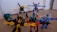 A group of colorful toy robots arranged on a wooden floor, featuring vibrant colors and intricate designs. Each robot is made of interlocking pieces in various poses, showcasing imaginative play. The scene captures a dynamic and playful moment, with toys spread out across the floor.