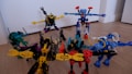 A group of colorful toy robots arranged on a wooden floor, featuring vibrant colors and intricate designs. Each robot is made of interlocking pieces in various poses, showcasing imaginative play. The scene captures a dynamic and playful moment, with toys spread out across the floor.