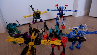 A group of colorful toy robots arranged on a wooden floor, featuring vibrant colors and intricate designs. Each robot is made of interlocking pieces in various poses, showcasing imaginative play. The scene captures a dynamic and playful moment, with toys spread out across the floor.