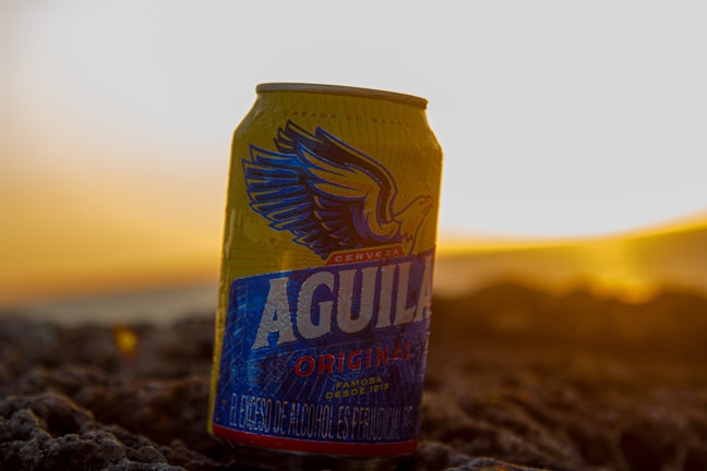 A can of Aguila beer is positioned on a rocky surface during a sunset. The eagle logo and branding are prominently displayed, with warm sunlight creating a golden glow in the background.