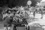 A bustling street scene captures a fruit vendor with a cart full of produce, including mangoes, pineapples, and melons. Several people are walking nearby, some wearing face masks. The background features trees providing shade and buses traveling along the road, with city buildings partly visible.