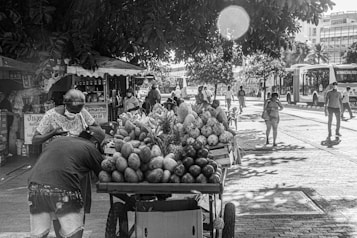 A bustling street scene captures a fruit vendor with a cart full of produce, including mangoes, pineapples, and melons. Several people are walking nearby, some wearing face masks. The background features trees providing shade and buses traveling along the road, with city buildings partly visible.