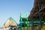 A beachfront scene with thatched-roof huts and elevated wooden structures painted in bright green. The sky is clear and blue, while the ocean is visible in the background.