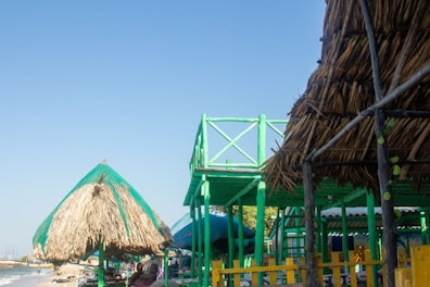 A beachfront scene with thatched-roof huts and elevated wooden structures painted in bright green. The sky is clear and blue, while the ocean is visible in the background.