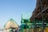 A beachfront scene with thatched-roof huts and elevated wooden structures painted in bright green. The sky is clear and blue, while the ocean is visible in the background.