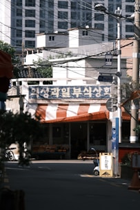 A street scene with a small grocery store displaying fresh produce under an orange and white striped canopy. In the background, there are several tall residential buildings. The foreground features trees and utility poles with numerous wires crossing overhead.