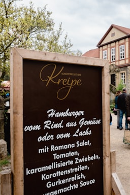 A rustic wooden sign with the name 'Kreipe' in elegant script at the top, advertising various burger options with ingredients like Romana salad and caramelized onions. It sits in front of a traditional building with a red-tiled roof and numerous windows. Surrounding greenery and a few people walking in the background are visible.