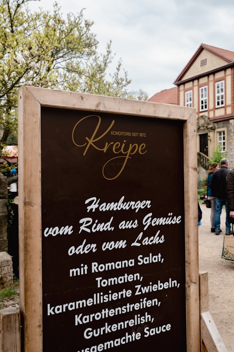 A rustic wooden sign with the name 'Kreipe' in elegant script at the top, advertising various burger options with ingredients like Romana salad and caramelized onions. It sits in front of a traditional building with a red-tiled roof and numerous windows. Surrounding greenery and a few people walking in the background are visible.