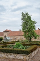 A well-maintained garden with neatly trimmed hedges and lush green plants, set against a backdrop of a large stone building with a red-tiled roof. A tall tree stands prominently next to the building, and there is a small greenhouse visible among the garden beds. The sky is overcast with gray clouds.