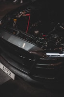 A close-up of a car's front end with the hood open, revealing internal components and wiring. The vehicle has a sleek, dark design with LED headlights and a customized license plate reading 'APEX'. The brand name 'Shelby' is visible on the bumper.