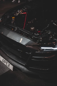 A close-up of a car's front end with the hood open, revealing internal components and wiring. The vehicle has a sleek, dark design with LED headlights and a customized license plate reading 'APEX'. The brand name 'Shelby' is visible on the bumper.