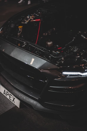 A close-up of a car's front end with the hood open, revealing internal components and wiring. The vehicle has a sleek, dark design with LED headlights and a customized license plate reading 'APEX'. The brand name 'Shelby' is visible on the bumper.