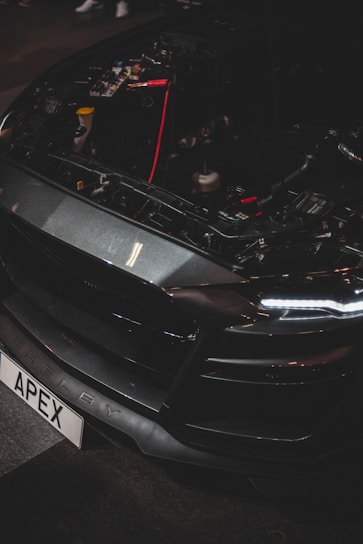 A close-up of a car's front end with the hood open, revealing internal components and wiring. The vehicle has a sleek, dark design with LED headlights and a customized license plate reading 'APEX'. The brand name 'Shelby' is visible on the bumper.