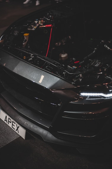 A close-up of a car's front end with the hood open, revealing internal components and wiring. The vehicle has a sleek, dark design with LED headlights and a customized license plate reading 'APEX'. The brand name 'Shelby' is visible on the bumper.