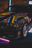 A stylish car with a vibrant, multicolored geometric pattern is parked indoors. The front bumper features a decal with '#POORBALTICS' and shiny alloy wheels. The lighting is dim, highlighting the colorful design.