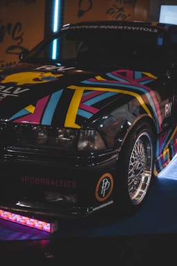 A stylish car with a vibrant, multicolored geometric pattern is parked indoors. The front bumper features a decal with '#POORBALTICS' and shiny alloy wheels. The lighting is dim, highlighting the colorful design.