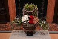 A lavish floral arrangement displayed in an ornate bronze vase. The arrangement includes white, red, and beige roses, along with other green foliage and smaller buds. Lavender and green foliage add texture to the bouquet. The background includes a tiled floor and a wooden wall, contributing to an elegant setting.