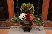 A lavish floral arrangement displayed in an ornate bronze vase. The arrangement includes white, red, and beige roses, along with other green foliage and smaller buds. Lavender and green foliage add texture to the bouquet. The background includes a tiled floor and a wooden wall, contributing to an elegant setting.