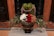 A lavish floral arrangement displayed in an ornate bronze vase. The arrangement includes white, red, and beige roses, along with other green foliage and smaller buds. Lavender and green foliage add texture to the bouquet. The background includes a tiled floor and a wooden wall, contributing to an elegant setting.
