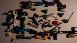 A collection of colorful plastic toy parts scattered on a surface. The parts include various shapes and sizes, such as connectors, rods, and small figures, predominantly made from interlocking pieces. The colors range from black, blue, green, yellow, white, red, to purple, with shadows cast by the parts adding to the scattered arrangement.
