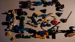 A collection of colorful plastic toy parts scattered on a surface. The parts include various shapes and sizes, such as connectors, rods, and small figures, predominantly made from interlocking pieces. The colors range from black, blue, green, yellow, white, red, to purple, with shadows cast by the parts adding to the scattered arrangement.