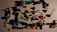 A collection of colorful plastic toy parts scattered on a surface. The parts include various shapes and sizes, such as connectors, rods, and small figures, predominantly made from interlocking pieces. The colors range from black, blue, green, yellow, white, red, to purple, with shadows cast by the parts adding to the scattered arrangement.