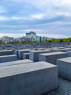 A series of large, rectangular concrete slabs arranged in a grid pattern. In the background, modern office buildings with glass facades rise against a cloudy sky. Sparse greenery intersperses the scene, providing a contrast to the geometric structures.