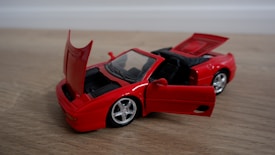 A red toy convertible car is placed on a wooden surface. The car's doors, trunk, and hood are open, revealing the interior details and engine space. The wheels are silver and shiny, and the overall condition of the toy is new.