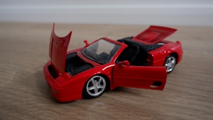A red toy convertible car is placed on a wooden surface. The car's doors, trunk, and hood are open, revealing the interior details and engine space. The wheels are silver and shiny, and the overall condition of the toy is new.