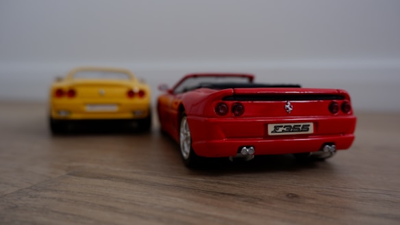 Two toy sports cars are positioned on a wooden surface. The foreground features a detailed model of a red convertible car, showcasing distinctive features such as taillights and a visible 'F355' license plate. The background includes a yellow car, slightly out of focus, contributing depth to the scene.
