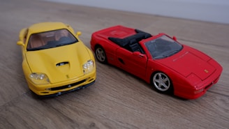 Two model sports cars are placed on a wooden surface. The car on the left is bright yellow with a sleek design and a closed roof. The car on the right is bright red and has a convertible, open-top design. Both cars have distinctive, sporty features and are highly detailed.