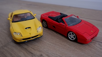 Two model sports cars are placed on a wooden surface. The car on the left is bright yellow with a sleek design and a closed roof. The car on the right is bright red and has a convertible, open-top design. Both cars have distinctive, sporty features and are highly detailed.