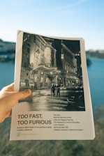 A hand holding a document with a black and white cover featuring a night scene of an urban setting with buildings and street lights. The title 'Too Fast, Too Furious' is prominently displayed, with text below indicating it is a bachelor's thesis from Lund University, focused on traffic's impact in Stockholm.