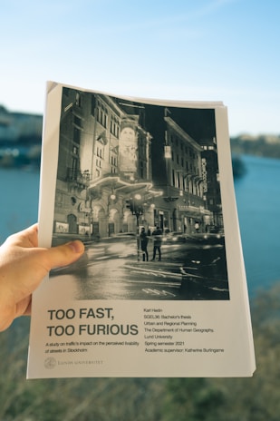 A hand holding a document with a black and white cover featuring a night scene of an urban setting with buildings and street lights. The title 'Too Fast, Too Furious' is prominently displayed, with text below indicating it is a bachelor's thesis from Lund University, focused on traffic's impact in Stockholm.