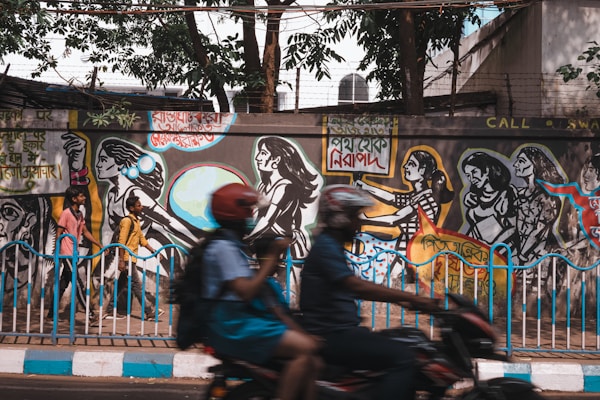 A vibrant street mural depicting a series of black and white figures in dynamic poses, painted against a background that includes colorful geometric shapes. Two people walk past the mural, capturing a blend of realism and art. The mural features text in what appears to be a regional script. The scene is lively with a blurred person riding a motorbike in the foreground.