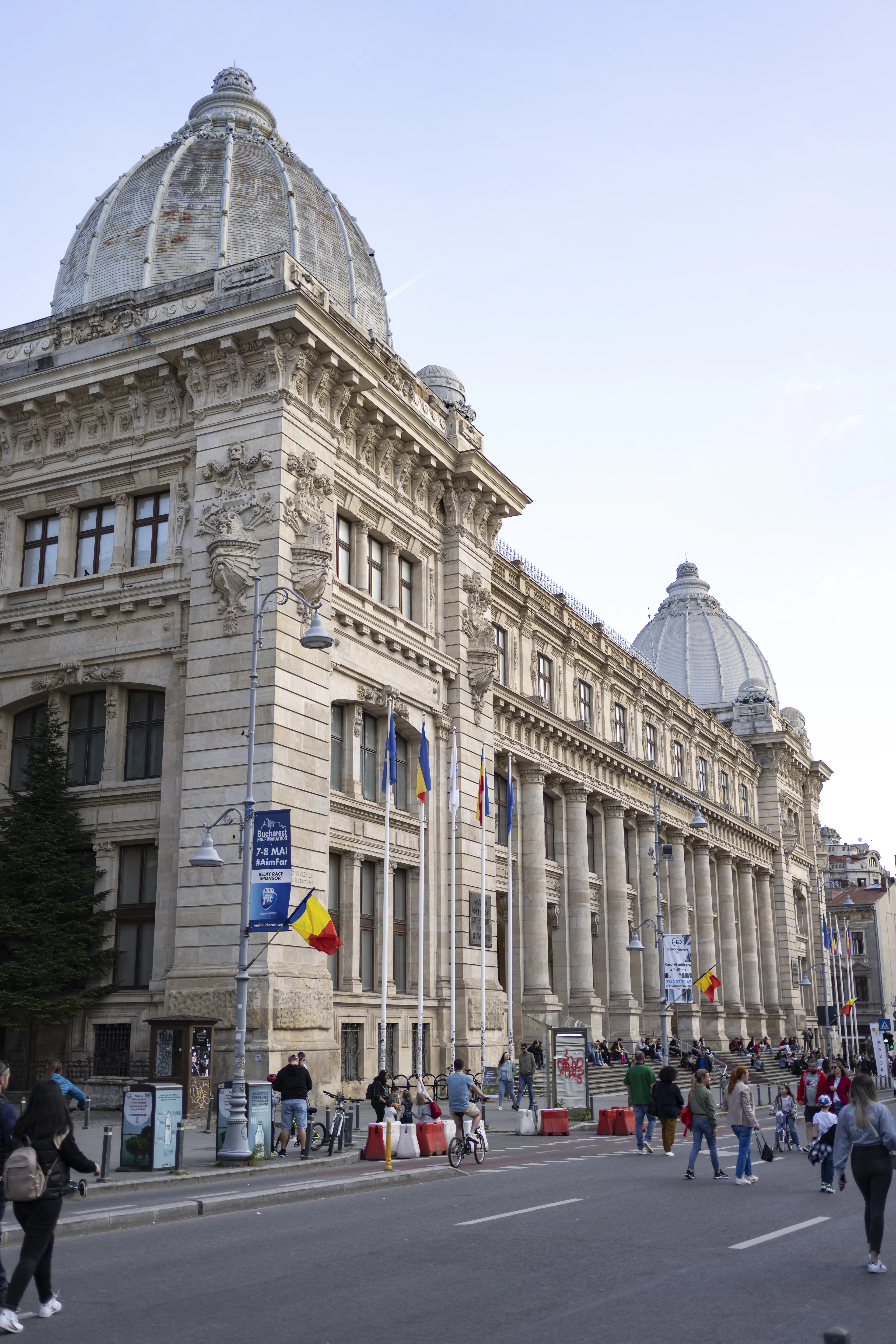 A domed Beaux-Arts building lines a Bucharest street as pedestrians pass beneath its columned facade.