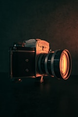 A vintage camera is prominently displayed against a dark background. The camera is metallic, with intricate dials and a large lens that emits a warm, orange glow. The lighting highlights the texture of the camera and creates a moody atmosphere.