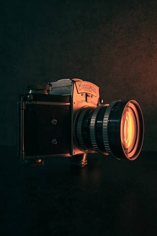 A vintage camera is prominently displayed against a dark background. The camera is metallic, with intricate dials and a large lens that emits a warm, orange glow. The lighting highlights the texture of the camera and creates a moody atmosphere.