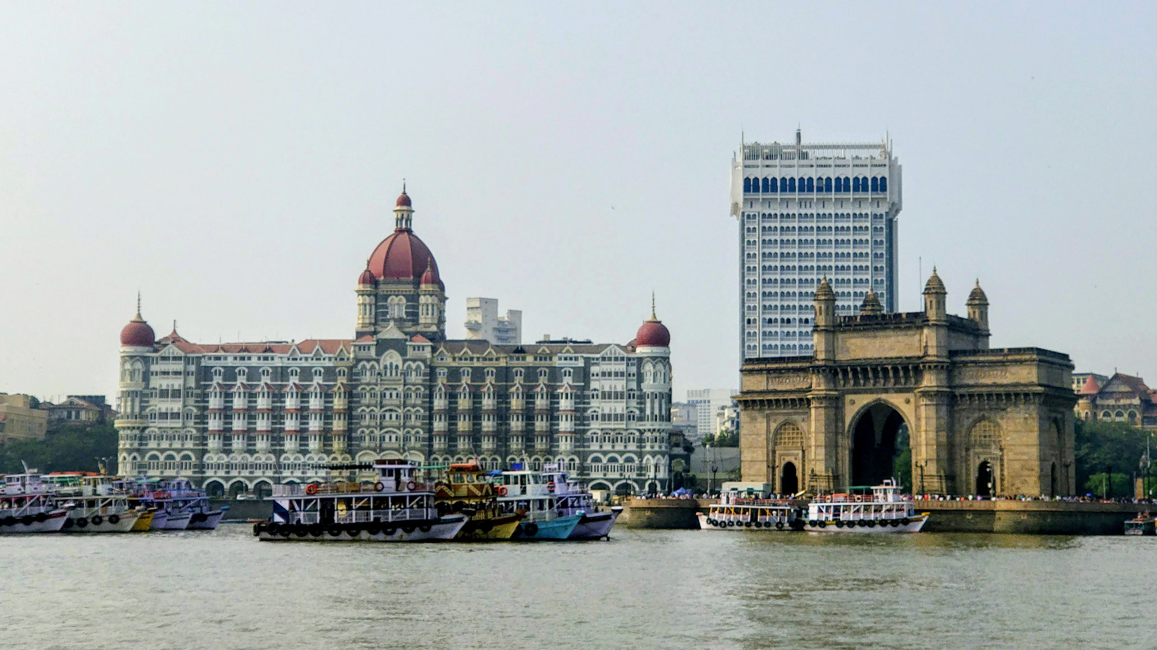 The domed Taj Mahal Palace Hotel and the Gateway of India arch seen across the harbor, with colorful tourist boats in the water.