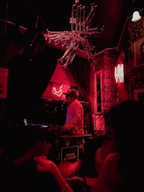 A DJ wearing headphones operates turntables in a dimly lit nightclub with vibrant red lighting. Decorative trumpets form a chandelier above, and the walls are adorned with various art and graffiti. Patrons, slightly blurred, dance in the foreground.