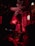 A DJ wearing headphones operates turntables in a dimly lit nightclub with vibrant red lighting. Decorative trumpets form a chandelier above, and the walls are adorned with various art and graffiti. Patrons, slightly blurred, dance in the foreground.