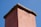 A close-up view of the top section of a brick chimney with a stone cap. The chimney is an earthy pink color and is set against a backdrop of a clear blue sky.