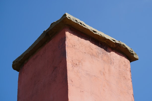 A close-up view of the top section of a brick chimney with a stone cap. The chimney is an earthy pink color and is set against a backdrop of a clear blue sky.