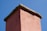 A close-up view of the top section of a brick chimney with a stone cap. The chimney is an earthy pink color and is set against a backdrop of a clear blue sky.