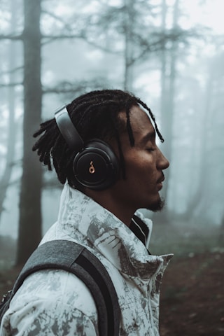 A man stands in a foggy forest with his eyes closed, wearing large black headphones and a white patterned jacket. He has a contemplative or relaxed expression as he seems to be listening to music.