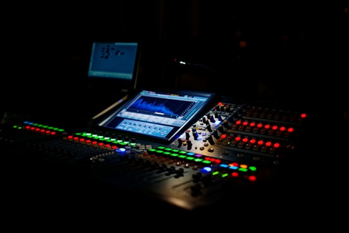 A dimly lit audio mixing console with illuminated buttons and displays. The central screen displays sound waveforms, and various knobs and sliders are visible. The atmosphere suggests a professional studio setting.