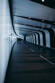 A modern, curved tunnel with a sleek design featuring alternating black and white panels along the walls and ceiling. The floor is dark with subtle geometric patterns, and there are several people walking through the tunnel, silhouetted against the illuminated panels.