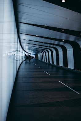 A modern, curved tunnel with a sleek design featuring alternating black and white panels along the walls and ceiling. The floor is dark with subtle geometric patterns, and there are several people walking through the tunnel, silhouetted against the illuminated panels.