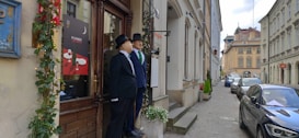 A narrow European street lined with historical buildings and parked cars. Two life-sized statues of men dressed in old-fashioned suits and hats stand at the entrance of a restaurant, which is adorned with green vines and a sign featuring wine glasses. The cobblestone sidewalk and classic architecture evoke a sense of timelessness.