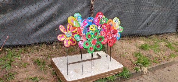 A group of colorful, flower-shaped pinwheels with intricate designs placed on metal stakes. These pinwheels are set on a white and brown rectangular base, situated outdoors on a patch of grass near a metal mesh fence.